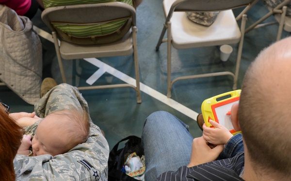 An overhead view of adults seated on folding chairs, including a person in U.S. Air Force ABU (Airman Battle Uniform) holding a sleeping infant. A bald man in a striped shirt sits nearby, with a young child holding a yellow toy drawing board. The setting appears to be an indoor gathering or waiting area with multiple folding chairs visible.