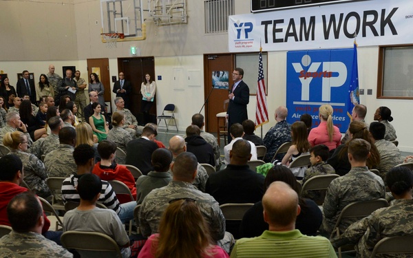 A large indoor gathering is taking place in what appears to be a military base gymnasium or community center. A speaker stands at a podium addressing a large audience composed predominantly of military personnel in camouflage uniforms, along with some civilians. Banners in the background reference 'TEAM WORK,' 'YP' (Youth Programs), 'Air Force Services,' and 'Boys &amp; Girls Clubs of America.'