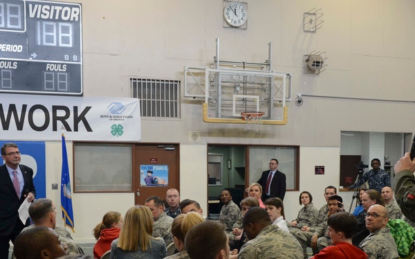 A formal gathering is taking place in a military base gymnasium, where a civilian official in a suit is addressing an audience of military personnel in camouflage uniforms and their families. A military officer in a flight suit stands to the right holding a microphone, likely facilitating a Q&amp;A session. The setting includes Air Force branding, a 'TEAM WORK' banner, Boys &amp; Girls Clubs of America signage, and American flags.