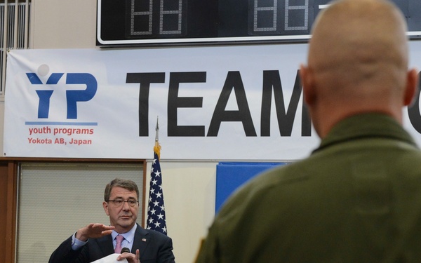 A civilian official in a suit and tie is addressing a group of military personnel at Yokota Air Base in Japan. The event appears to be a formal briefing or town hall meeting held in what looks like a gymnasium or community center. A banner for 'Youth Programs Yokota AB, Japan' and a partial 'TEAM' banner are visible in the background.