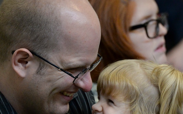 A bald man wearing black-rimmed glasses and a dark pinstripe shirt shares a joyful, close moment with a young blonde child wearing a denim jacket. Both are smiling broadly and facing each other in an affectionate interaction. A red-haired woman wearing glasses is visible in the background.