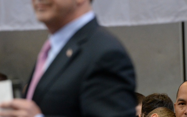 A formal event or ceremony is taking place, likely a public meeting, graduation, or civic gathering. In the foreground, a man in a suit and tie holds a child who is resting their head on his shoulder in an affectionate embrace. In the background, a blurred figure in a dark suit with a pink tie appears to be speaking or presenting at the event.