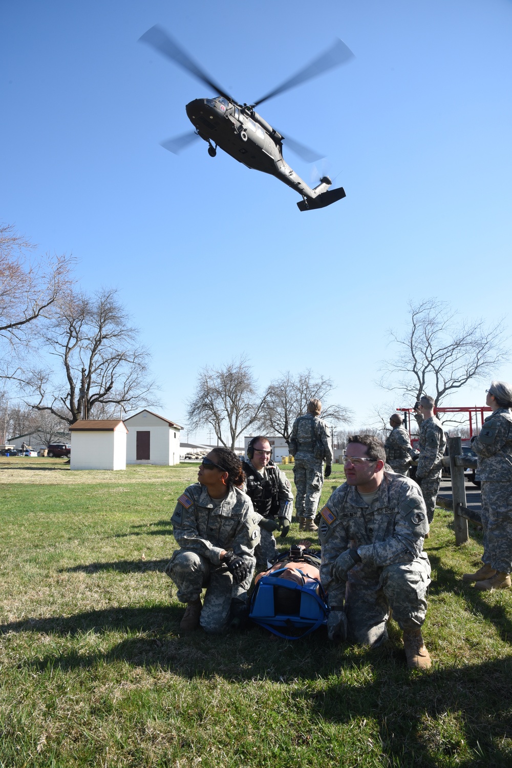 A military medical evacuation (MEDEVAC) training exercise is underway on a grassy field. U.S. Army soldiers in ACU (Army Combat Uniform) are attending to a simulated casualty on a blue litter/stretcher while a UH-60 Black Hawk helicopter hovers overhead. The scene depicts a realistic CASEVAC/MEDEVAC drill with multiple personnel in coordinated roles.