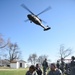 A military medical evacuation (MEDEVAC) training exercise is underway on a grassy field. U.S. Army soldiers in ACU (Army Combat Uniform) are attending to a simulated casualty on a blue litter/stretcher while a UH-60 Black Hawk helicopter hovers overhead. The scene depicts a realistic CASEVAC/MEDEVAC drill with multiple personnel in coordinated roles.