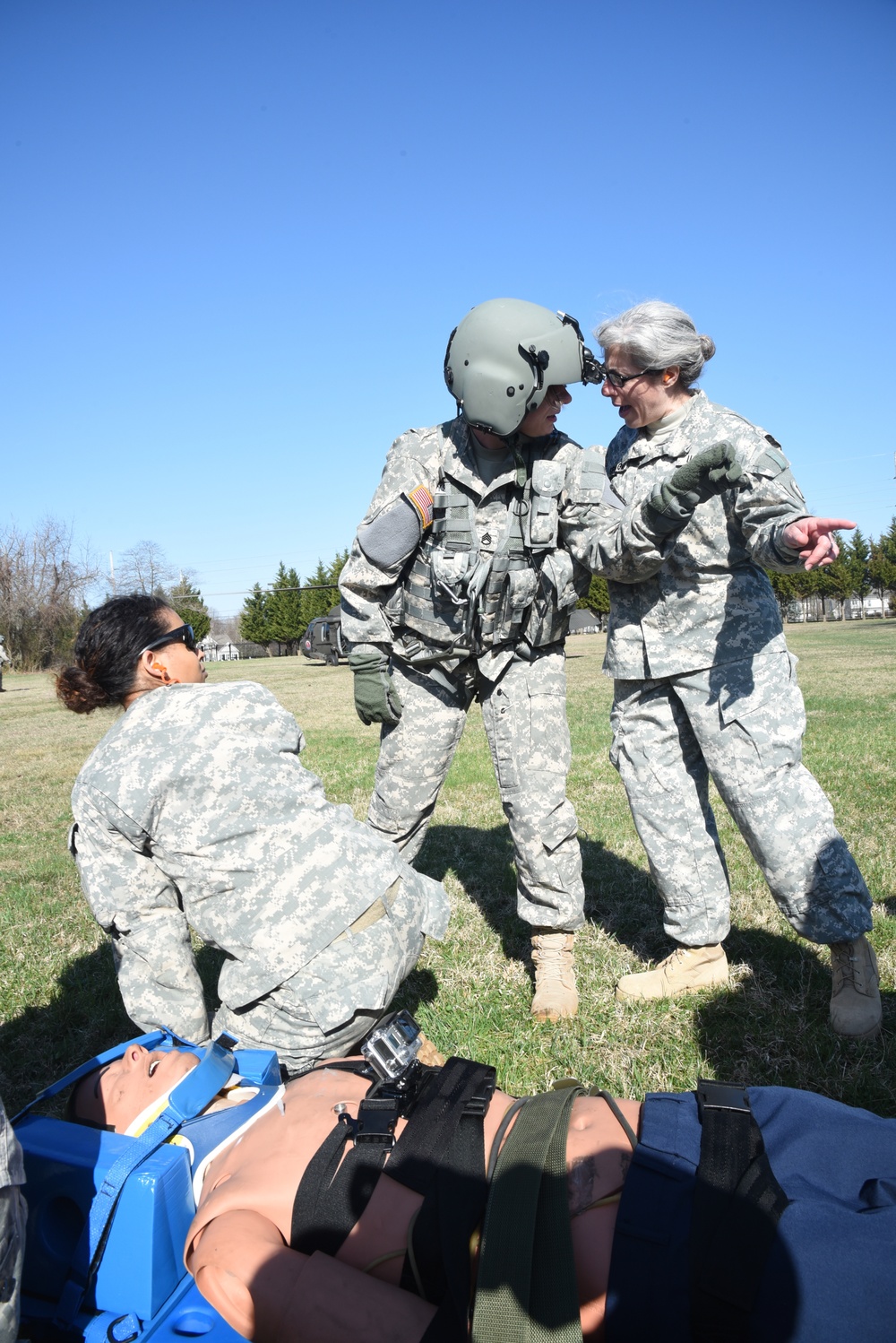 Three U.S. Army soldiers in ACU (Army Combat Uniform) pattern uniforms are engaged in what appears to be a medical training exercise outdoors on a grassy field. One soldier wearing a flight helmet and tactical vest stands in the center, while a senior soldier (with gray hair) appears to be providing instruction or debriefing. A third soldier kneels beside a simulated casualty strapped to a blue litter/stretcher on the ground.