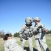 Three U.S. Army soldiers in ACU (Army Combat Uniform) pattern uniforms are engaged in what appears to be a medical training exercise outdoors on a grassy field. One soldier wearing a flight helmet and tactical vest stands in the center, while a senior soldier (with gray hair) appears to be providing instruction or debriefing. A third soldier kneels beside a simulated casualty strapped to a blue litter/stretcher on the ground.