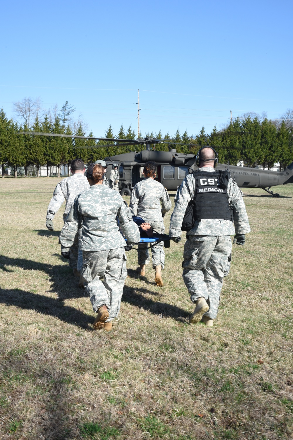 Military personnel in U.S. Army ACU (Army Combat Uniform) are carrying a patient on a stretcher toward a UH-60 Black Hawk helicopter for a medical evacuation (MEDEVAC). One individual is wearing a black tactical vest labeled 'CST MEDICAL' and communications headset. This appears to be a training exercise or real casualty evacuation scenario on an open grassy field.