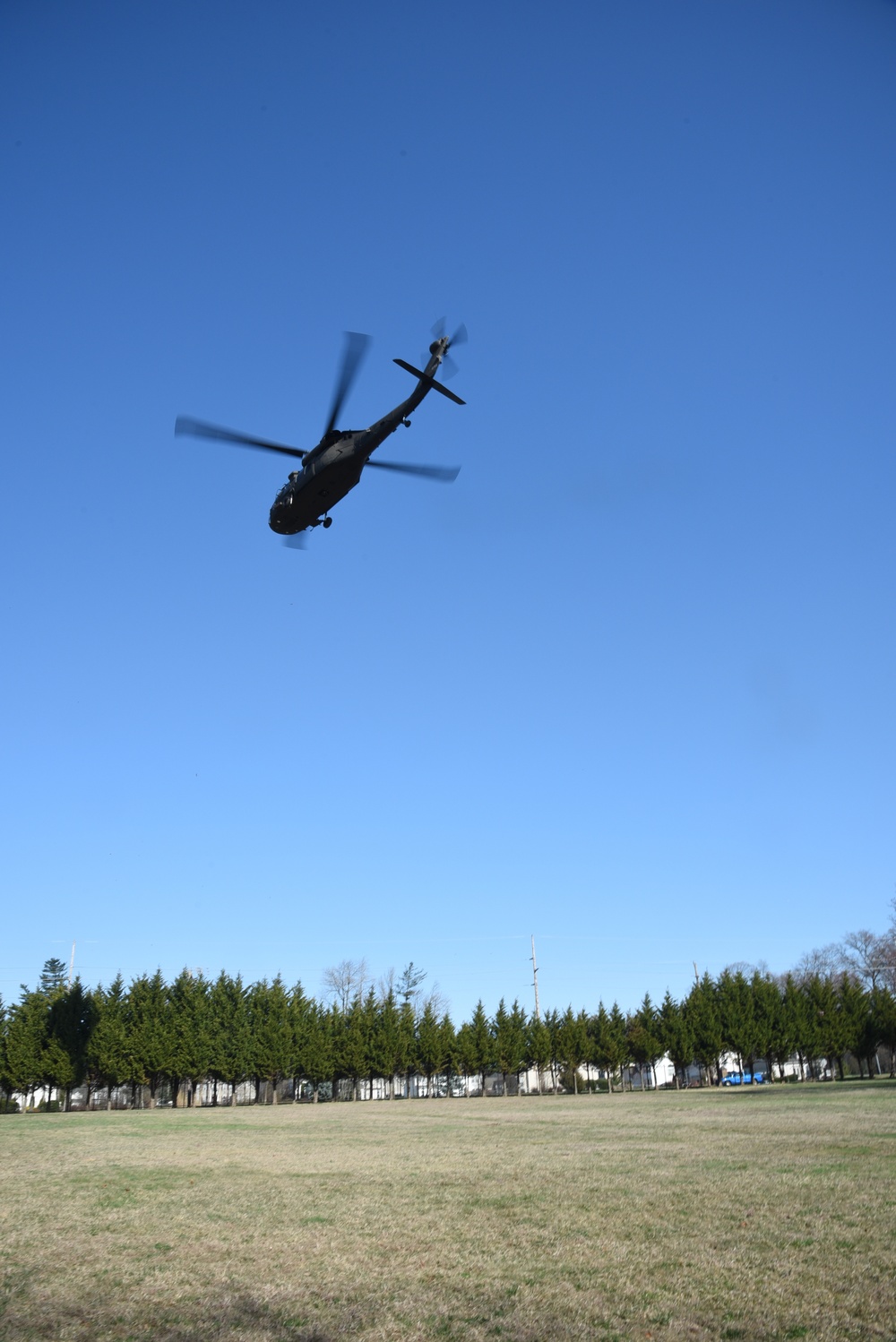 A military helicopter, consistent with a UH-60 Black Hawk, is captured in flight from a low-angle perspective against a clear blue sky. The helicopter appears to be flying at low altitude over an open grassy field bordered by a row of evergreen trees. Buildings and a utility pole are visible in the background beyond the tree line.