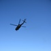 A military helicopter, consistent with a UH-60 Black Hawk, is captured in flight from a low-angle perspective against a clear blue sky. The helicopter appears to be flying at low altitude over an open grassy field bordered by a row of evergreen trees. Buildings and a utility pole are visible in the background beyond the tree line.