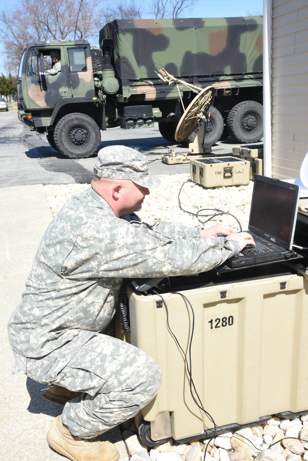 A U.S. Army soldier in ACU (Army Combat Uniform) is crouching and operating a ruggedized laptop computer placed on top of a tan military equipment case marked '1280'. Behind him is a portable satellite dish antenna and additional military cases, with a camouflage-painted military truck (LMTV or similar) visible in the background. The setting appears to be a field communications or command post setup in a paved area near a building.