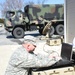 A U.S. Army soldier in ACU (Army Combat Uniform) is crouching and operating a ruggedized laptop computer placed on top of a tan military equipment case marked '1280'. Behind him is a portable satellite dish antenna and additional military cases, with a camouflage-painted military truck (LMTV or similar) visible in the background. The setting appears to be a field communications or command post setup in a paved area near a building.