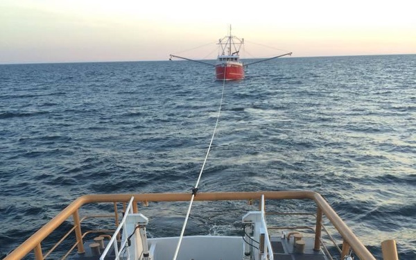 A U.S. Coast Guard vessel is actively towing a red commercial fishing vessel across open ocean waters. A Coast Guard crew member in an orange life vest stands on the stern deck monitoring the tow line connected to the distressed fishing boat in the distance. The scene appears to be taking place at dusk or dawn based on the soft light on the horizon.