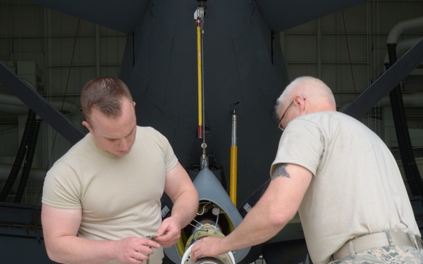 Two U.S. Air Force personnel in ABU (Airman Battle Uniform) camouflage are working on a component in what appears to be an aircraft hangar. They are handling what appears to be a munition or ordnance nose cone/fuze assembly in front of a large dark-colored aircraft. A yellow safety stand and hazard markings are visible in the background.