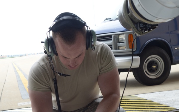 A military airman in US Air Force camouflage uniform and hearing protection is crouched on an airfield tarmac, working with a small electronic/electrical junction box device with multiple cables. He appears to be performing maintenance or testing operations on flight line equipment. A blue and silver Chevrolet van is visible in the background on the tarmac.