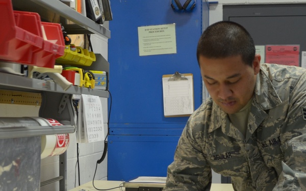 A U.S. Air Force airman in ABU (Airman Battle Uniform) is writing on paperwork at a work counter, with a FedEx shipping label visible beneath his hand. The setting appears to be a military TMO (Traffic Management Office) or shipping/logistics facility. The airman's name tape reads 'AGUILAR' and rank insignia indicates Staff Sergeant.