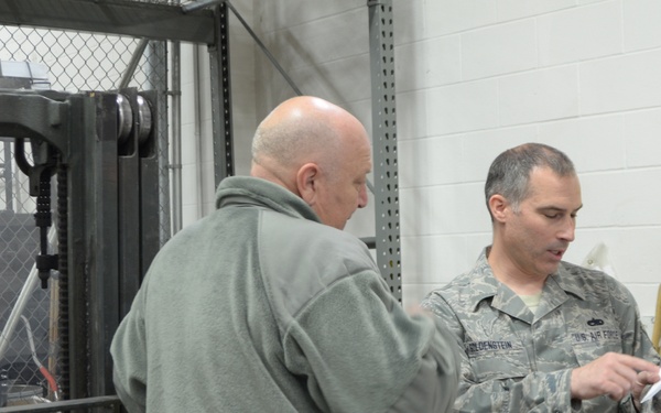 Two individuals are standing in what appears to be a military warehouse or storage facility. One person is wearing a US Air Force camouflage uniform and is reviewing a document, while the other is in civilian clothing. They appear to be conducting an inspection or inventory review near a forklift and a yellow flammable materials cabinet.