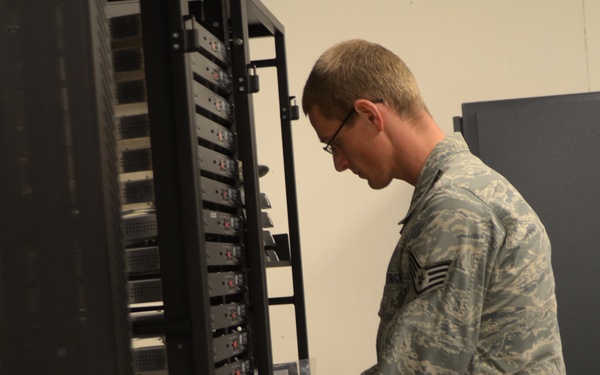 A U.S. Air Force enlisted member in Airman Battle Uniform (ABU) is working on a server or equipment rack in what appears to be a data center or communications room. The individual is examining or installing rack-mounted equipment units while referencing a document or manual. An open cardboard box is visible nearby, suggesting new equipment installation or maintenance work.