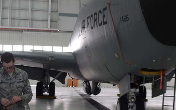 A U.S. Air Force airman in ABU (Airman Battle Uniform) is performing maintenance work on a military aircraft inside a hangar. The aircraft, marked 'U.S. AIR FORCE' with tail number 1466, appears to be a large jet aircraft undergoing inspection or servicing. The airman is working near yellow Pelican equipment cases and various ground support equipment.