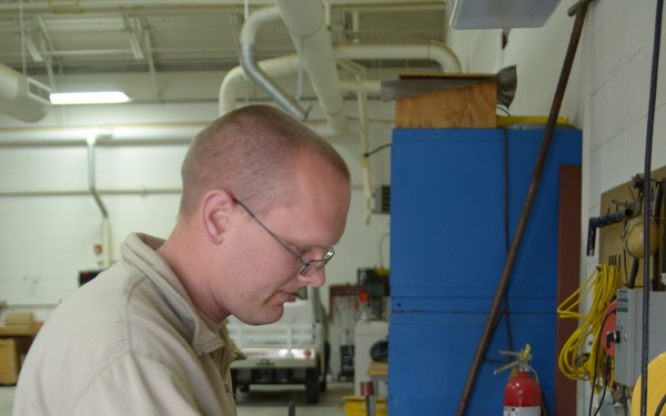 A man wearing US Air Force digital camouflage (ABU) pants and a tan fleece jacket is working on a large industrial valve or mechanical component at a workbench in what appears to be a maintenance shop or hangar. He is wearing glasses and a ring, and is focused on assembling or inspecting the gray flanged component. The background shows a well-equipped workshop with tools, a fire extinguisher, extension cords, and a vehicle visible in the distance.