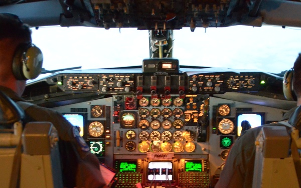 Two pilots are seated in the cockpit of a large aircraft, viewed from behind. The flight deck features an extensive array of analog gauges, digital displays, and illuminated control panels consistent with a military or older commercial aircraft. The cockpit is lit with a mix of ambient daylight from the windscreen and instrument panel lighting, including green and red illumination.