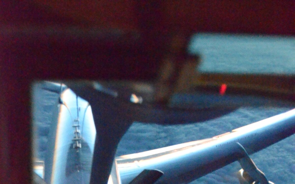 This image shows an aerial refueling operation viewed from the boom operator's station of a tanker aircraft. A large multi-engine aircraft, consistent with a B-52 Stratofortress, is positioned below and behind the tanker, with the refueling boom extended and connected to the receiver aircraft's refueling receptacle. The image is taken through the boom operator's window, with red instrument lighting visible in the foreground and a cloud layer visible below.