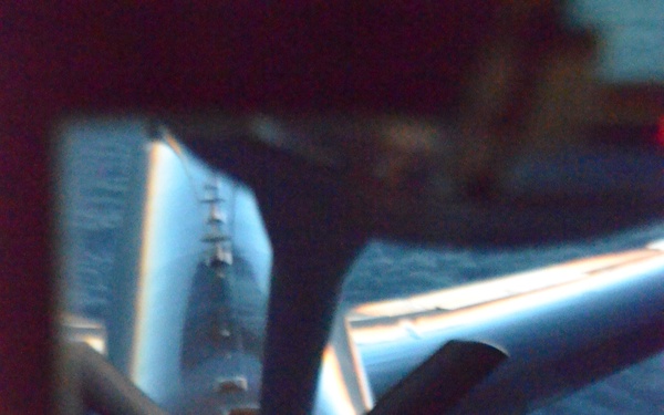 The image shows an aerial refueling operation captured from the boom operator's position aboard a tanker aircraft, looking down through the boom operator's window at a receiver aircraft below. The receiver aircraft, which appears to be a large military or commercial-type aircraft, is visible with its refueling receptacle illuminated by orange/yellow lights. The ocean or large body of water is visible in the background, suggesting the operation is taking place at altitude over water.