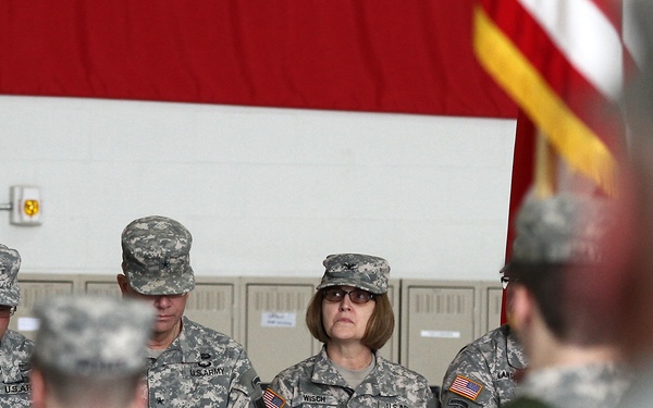 A group of U.S. Army soldiers in ACU (Army Combat Uniform) digital camouflage are seated at what appears to be a formal military ceremony or briefing. A female soldier with the name tape 'WISCH' is prominently visible in the center, wearing glasses and a colonel's eagle insignia on her patrol cap. An American flag and a red curtain/drape are visible in the background.