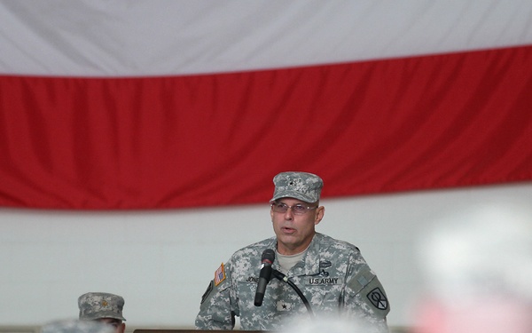 A U.S. Army officer, wearing ACU (Army Combat Uniform) with what appears to be a general officer rank insignia on his cap, is speaking at a podium with a microphone. He is addressing an audience of uniformed soldiers seated in the foreground. The event appears to be a formal military ceremony or briefing, with a large red and white draped flag and a unit guidon visible in the background.
