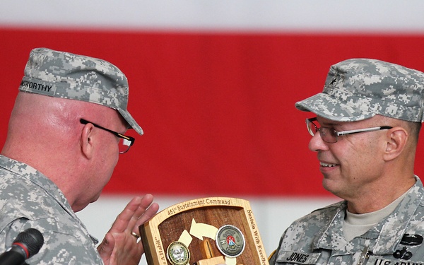 Two U.S. Army soldiers in ACU (Army Combat Uniform) are engaged in a military award or recognition ceremony. The soldier on the right, identified by his name tape as 'JONES' and wearing a Brigadier General rank insignia (one star) along with a medal, is receiving or presenting a wooden shield plaque. The plaque is inscribed with '451st Sustainment Command' and references 'BG Kenneth D. Jones' for leadership and dedication.