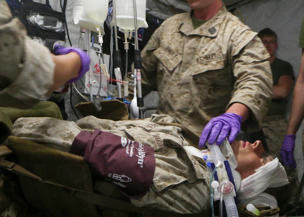 A military medical treatment scene showing a casualty in MARPAT camouflage uniform receiving emergency care on a litter. Medical personnel wearing purple nitrile gloves are administering treatment including IV fluids and airway management with an endotracheal tube. The setting appears to be a forward medical facility or field hospital tent.