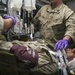 A military medical treatment scene showing a casualty in MARPAT camouflage uniform receiving emergency care on a litter. Medical personnel wearing purple nitrile gloves are administering treatment including IV fluids and airway management with an endotracheal tube. The setting appears to be a forward medical facility or field hospital tent.