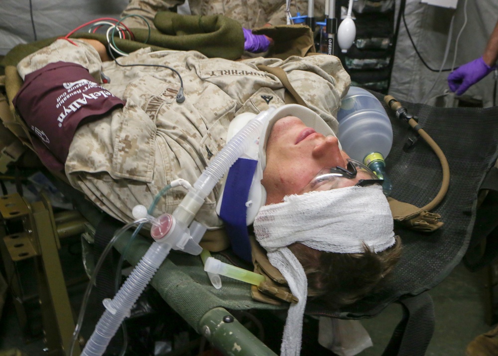 A U.S. Marine in desert camouflage uniform is receiving emergency medical treatment, lying on a field stretcher or litter. The patient has multiple medical devices attached including a breathing mask, cervical collar, head bandaging, blood pressure cuff, and monitoring leads. Medical personnel wearing purple nitrile gloves are visible attending to the patient in what appears to be a field medical facility or combat support hospital.