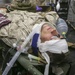 A U.S. Marine in desert camouflage uniform is receiving emergency medical treatment, lying on a field stretcher or litter. The patient has multiple medical devices attached including a breathing mask, cervical collar, head bandaging, blood pressure cuff, and monitoring leads. Medical personnel wearing purple nitrile gloves are visible attending to the patient in what appears to be a field medical facility or combat support hospital.