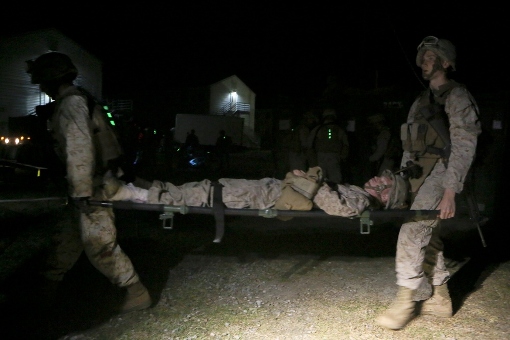 A nighttime military scene showing two soldiers in desert camouflage uniforms carrying a fellow soldier on a stretcher. Additional personnel in full combat gear are visible in the background near illuminated structures. The scene appears to be a training exercise or casualty evacuation operation.