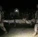 A nighttime military scene showing two soldiers in desert camouflage uniforms carrying a fellow soldier on a stretcher. Additional personnel in full combat gear are visible in the background near illuminated structures. The scene appears to be a training exercise or casualty evacuation operation.