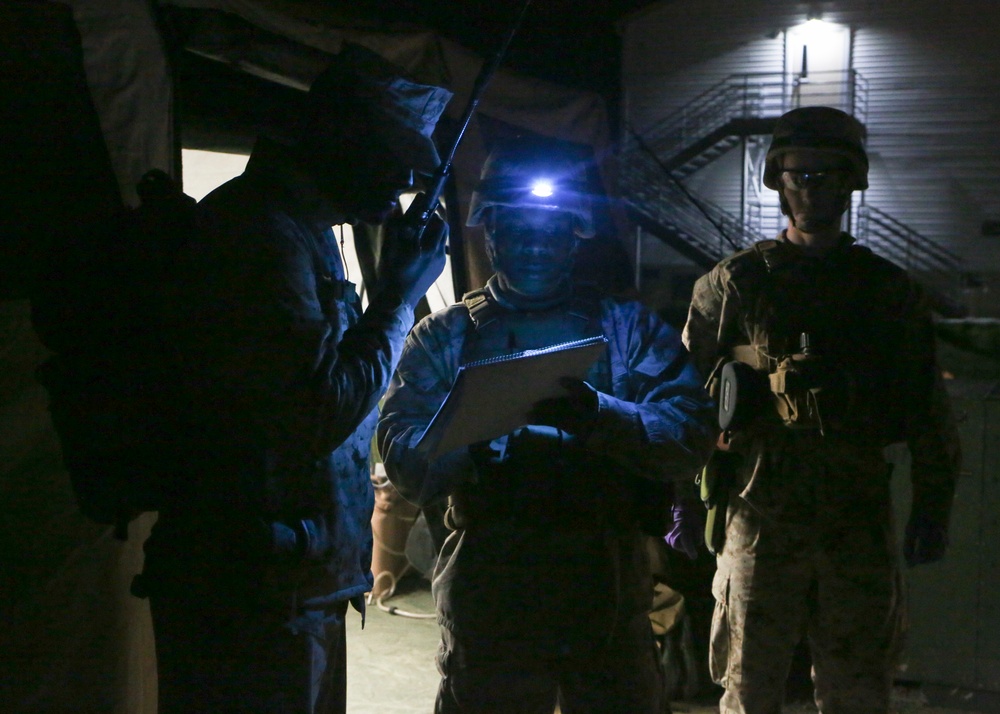A nighttime military operation scene showing three soldiers in combat gear operating in low-light conditions. One soldier is using a radio while another illuminates a notepad or clipboard with a headlamp mounted on their helmet. A third soldier stands in the background wearing a helmet and tactical vest.