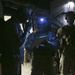 A nighttime military operation scene showing three soldiers in combat gear operating in low-light conditions. One soldier is using a radio while another illuminates a notepad or clipboard with a headlamp mounted on their helmet. A third soldier stands in the background wearing a helmet and tactical vest.