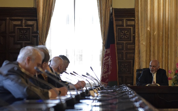 A formal meeting is taking place in an ornate conference room around a long polished table. One individual sits at the head of the table facing several attendees seated along one side. An Afghan national flag is prominently displayed in the background near a large window with gold curtains.