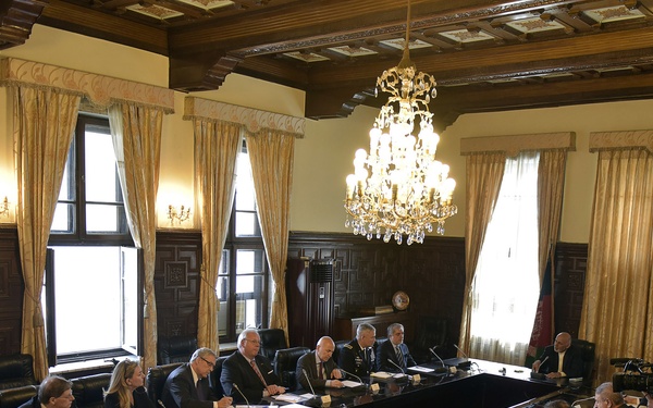 A formal high-level meeting is taking place in an ornate conference room with wood-paneled walls and a crystal chandelier. Two delegations are seated across from each other at a large rectangular conference table equipped with microphones and name placards. A photographer or videographer is visible on the right side documenting the proceedings.