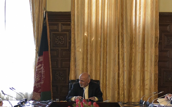 A formal high-level meeting is taking place in an ornate conference room, with a central figure seated at the head of a large rectangular table flanked by two delegations on either side. An Afghan national flag is prominently displayed behind the central figure, suggesting this may be an official government meeting in Afghanistan. The setting, decor, and arrangement indicate a diplomatic or governmental summit.