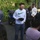 1-160th SOAR (A) feeds homeless, malnourished A group of people are gathered outdoors in what appears to be a parking area or driveway, with several individuals bowing their heads in what looks like a prayer or moment of reflection. A man in a white Nike long-sleeve shirt stands centrally, holding a dark cap, appearing to lead the gathering. Trees with green foliage and a motorcycle with a top box are visible in the background.