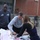 1-160th SOAR (A) feeds homeless, malnourished Three people are gathered around a makeshift outdoor table covered with bags, boxes, and food containers in what appears to be a parking area or alley beside a brick building. Two individuals are leaning over the table actively handling items, while a third person stands and observes. The setting and arrangement of food items suggest a food distribution or community meal activity.