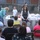 1-160th SOAR (A) feeds homeless, malnourished An outdoor community gathering taking place in a parking lot or alleyway adjacent to a brick building. A woman in a gray vest appears to be leading or addressing a group of people seated in folding chairs arranged in a semicircle. A folding table behind her holds numerous white plastic bags, suggesting a food or supply distribution event.