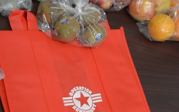 The image shows a red reusable tote bag branded with 'Operation Homefront Central Midwest' lying on a dark wooden table. Surrounding the bag are various fresh fruits and vegetables packaged in clear plastic bags, including kiwis, oranges, a pineapple, and green vegetables. A partially visible document labeled 'Healthy Garden' is visible in the upper right corner.