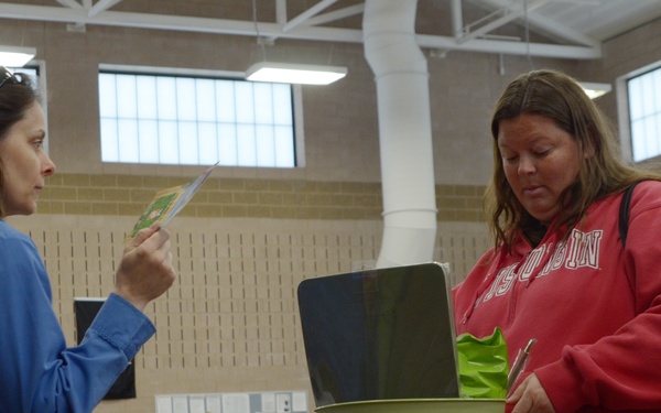 Two women are interacting at a booth during what appears to be a health fair or community expo held in a large indoor facility. One woman in a blue top is reviewing a pamphlet or card, while another in a red Wisconsin sweatshirt examines materials at the table. The background shows additional booths, attendees, and informational displays throughout the venue.