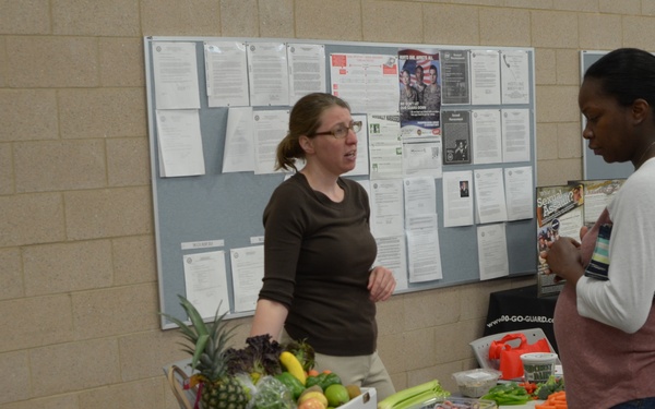 Two women are engaged in conversation at what appears to be a health or nutrition fair booth. A table between them displays a box of fresh fruits and vegetables including a pineapple, bananas, and various produce. The setting appears to be a gymnasium or community center with a bulletin board visible in the background.