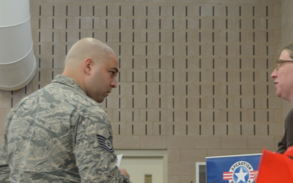 A U.S. Air Force service member in ABU (Airman Battle Uniform) camouflage is conversing with a civilian woman at what appears to be an Operation Homefront event. A table with fresh fruits and vegetables is visible between them, suggesting a food assistance or community support distribution event. A blue Operation Homefront banner is visible in the background.