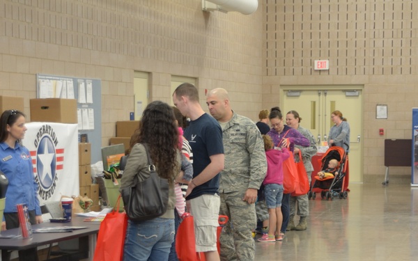 The image shows a military family support event, likely an 'Operation Homefront' distribution or resource fair held in a large indoor facility. Military personnel in camouflage uniforms and their families are lined up at tables staffed by volunteers in blue shirts. Participants are receiving red tote bags and other items, with cardboard boxes stacked in the background suggesting donated goods.