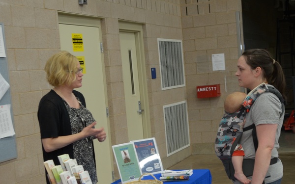 An indoor event or fair where a representative stands behind a blue-tablecloth booth, speaking with a woman who is carrying a baby in a front-facing carrier. The booth displays brochures, pens, a wicker basket, and materials bearing what appears to be a 'Hill Physicians' or similar healthcare/community organization logo. The setting appears to be a large warehouse or community center facility.