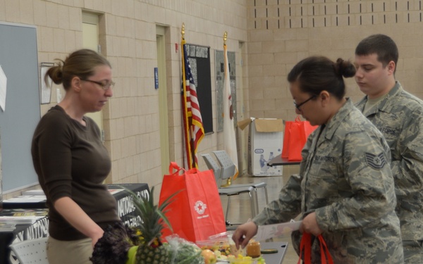 The image shows a military wellness or resource fair taking place in what appears to be a large indoor facility, likely a National Guard armory or military building. Two service members in Air Force camouflage uniforms (ACUs) are interacting with a civilian woman at a table displaying fresh fruits and informational materials. Red tote bags and National Guard branding are visible throughout the scene.